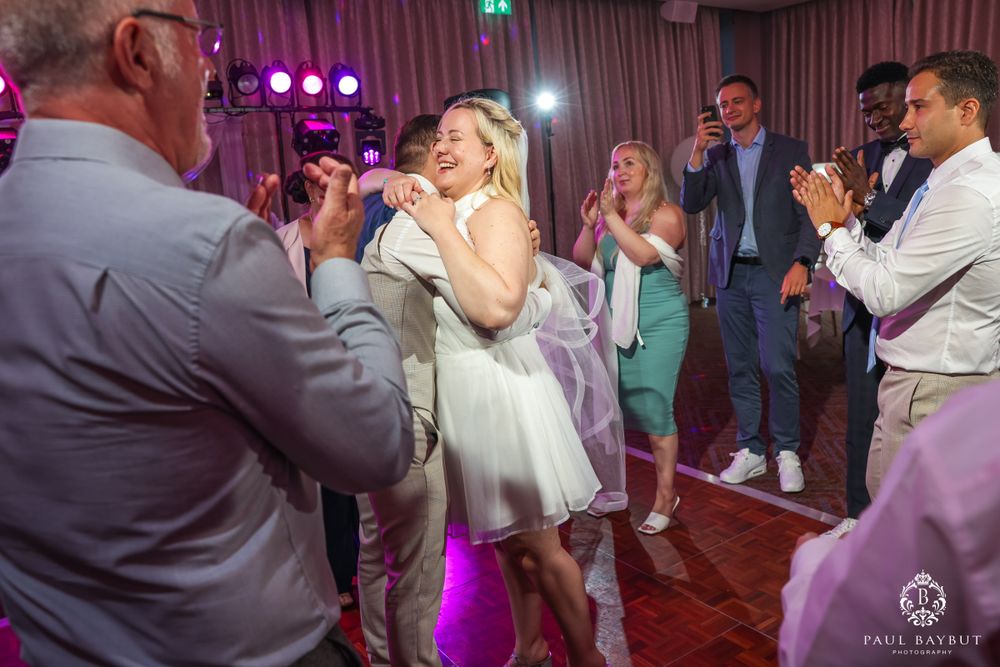 Wedding guests dancing and celebrating on a wooden dance floor under purple lighting.