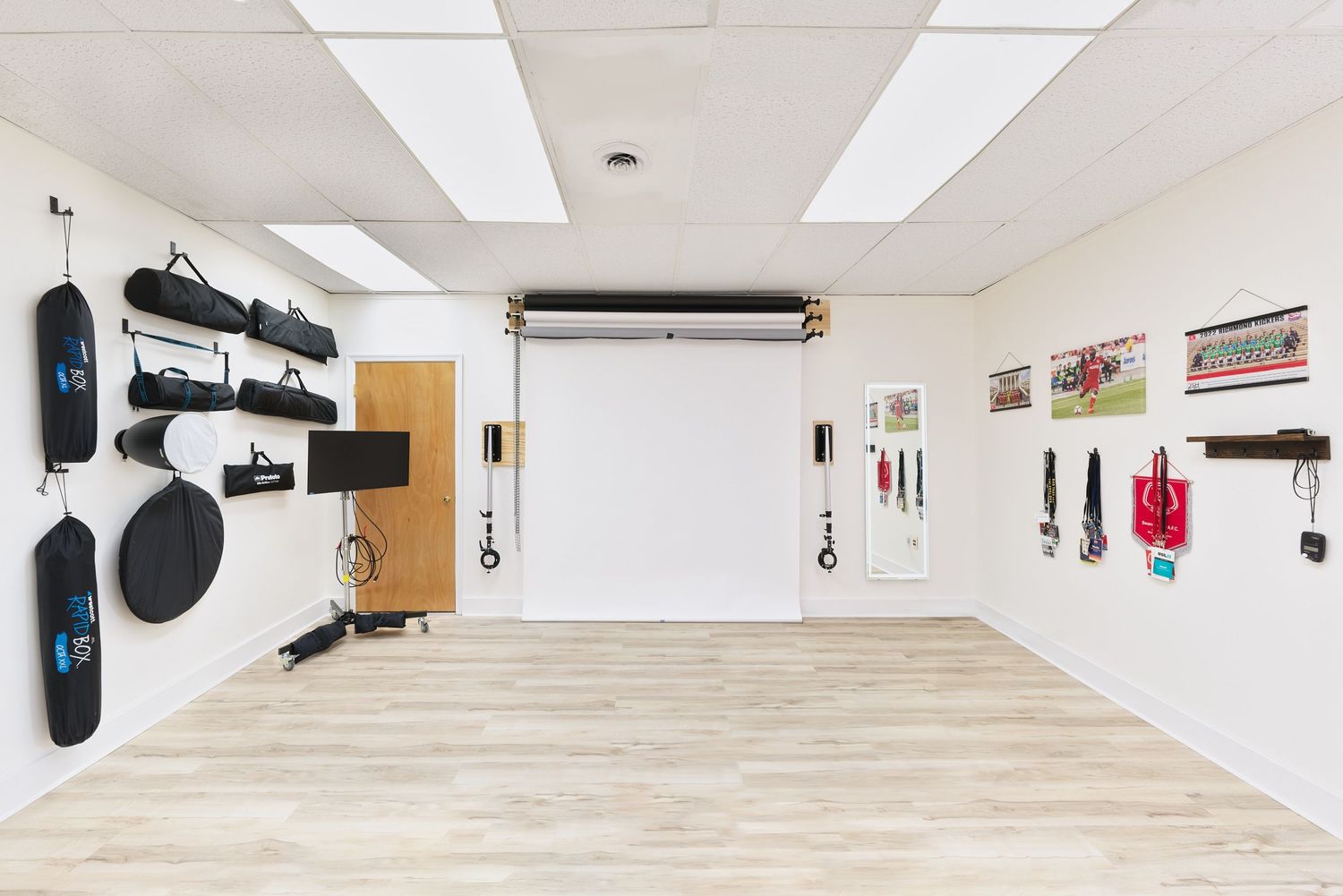 Empty photography studio with white backdrop, light wood flooring and photography equipment mounted on walls.