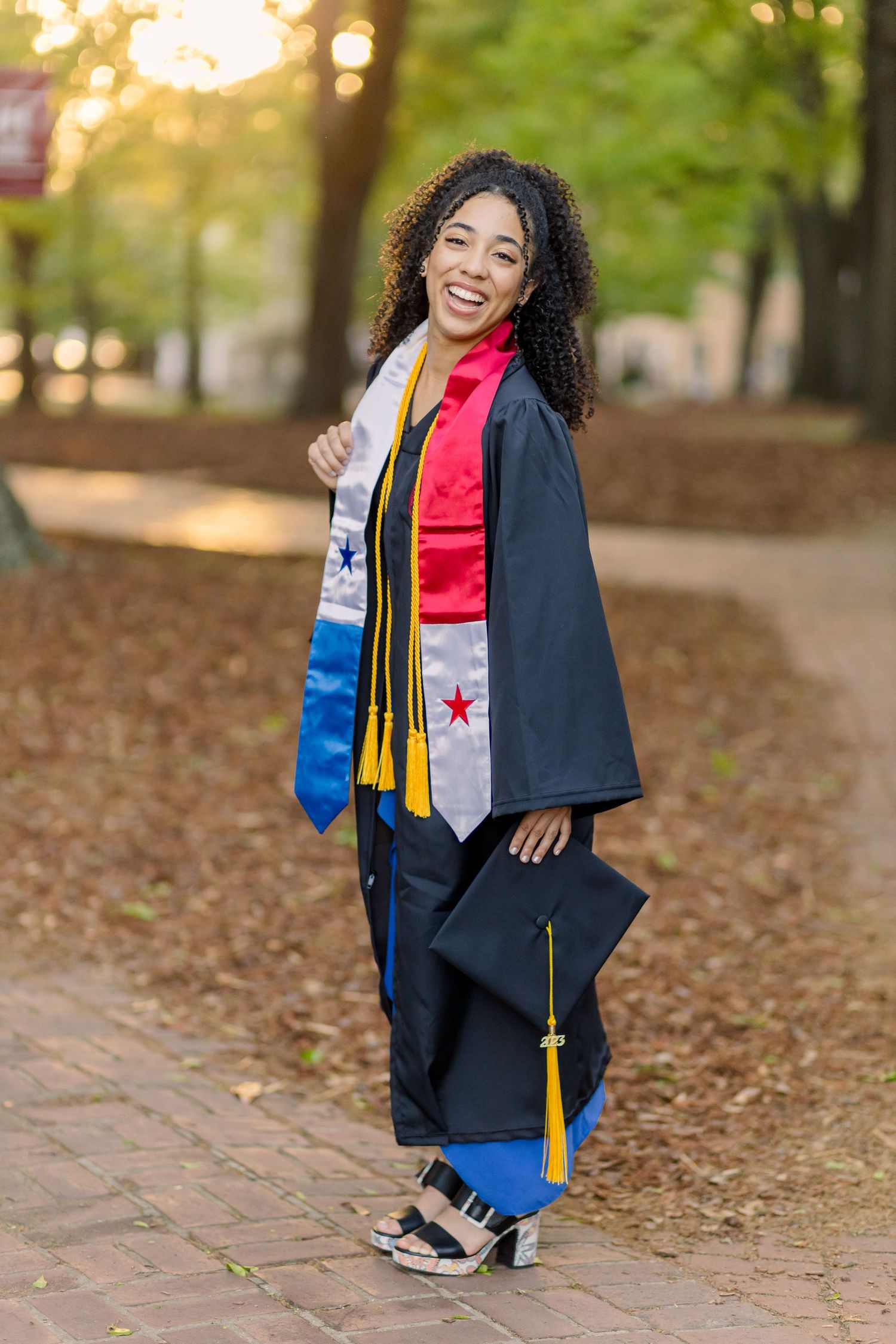 Graduate in navy blue cap and gown with colorful stoles poses outdoors during sunset on campus path.