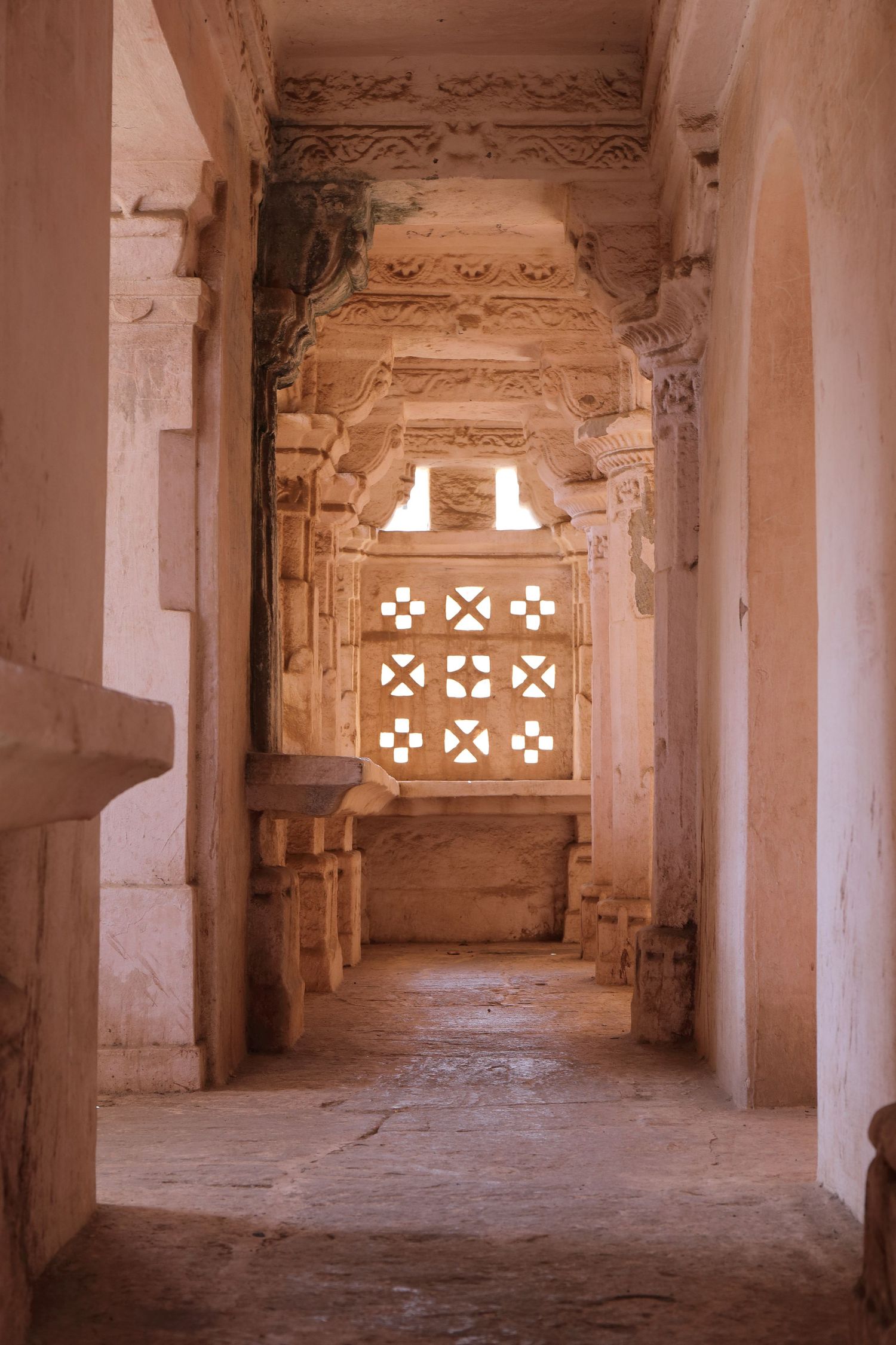 Ancient stone corridor with ornate ceiling details and geometric window patterns letting in natural light.