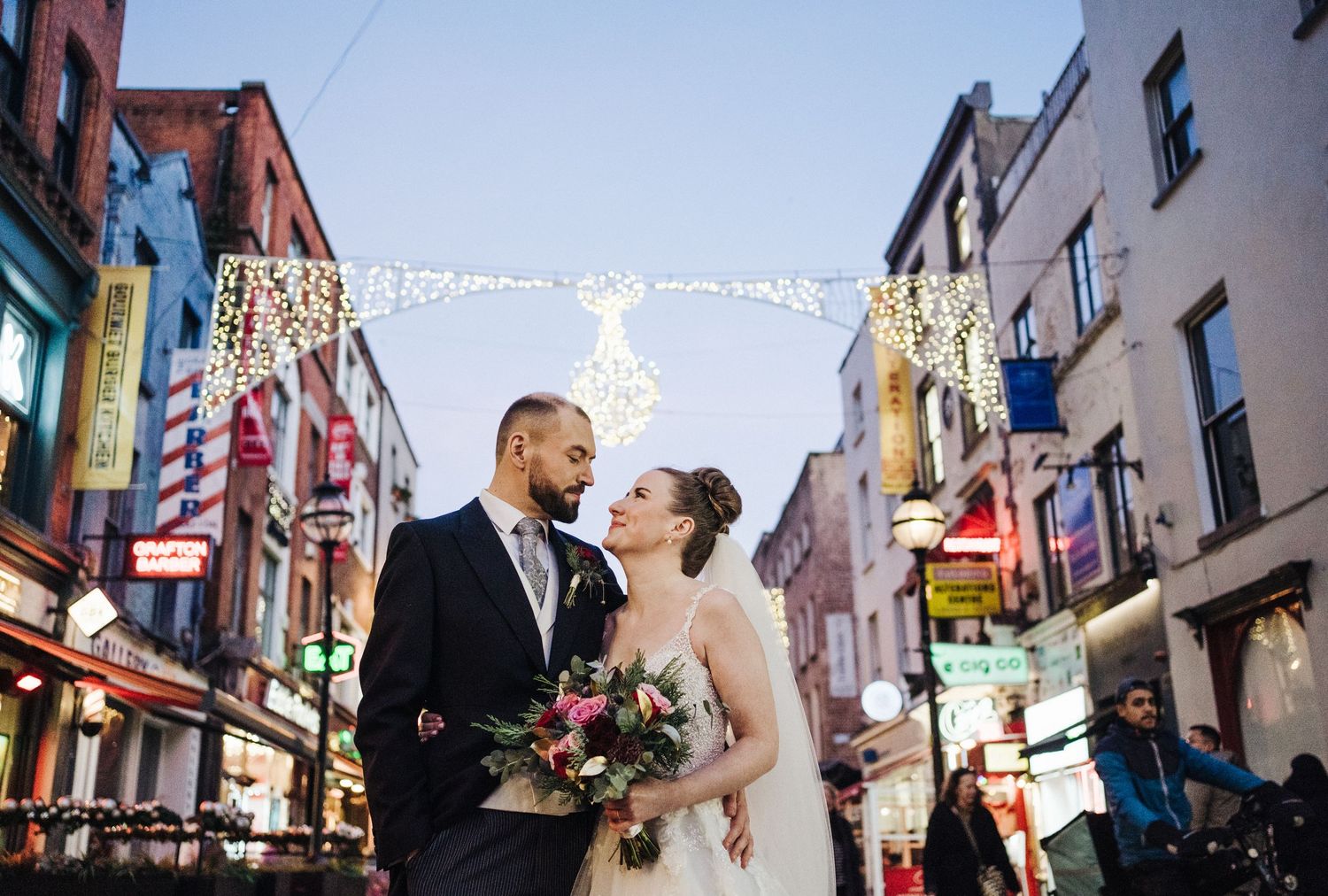 Newlyweds share intimate moment on city street with festive lights overhead during evening celebration.