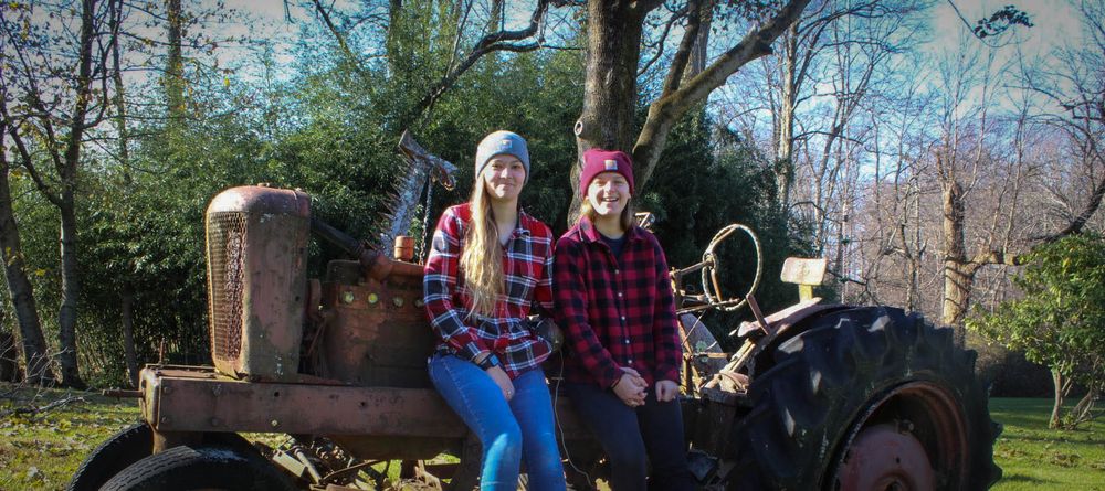 Two people in matching plaid shirts pose on a vintage tractor in a wooded setting.