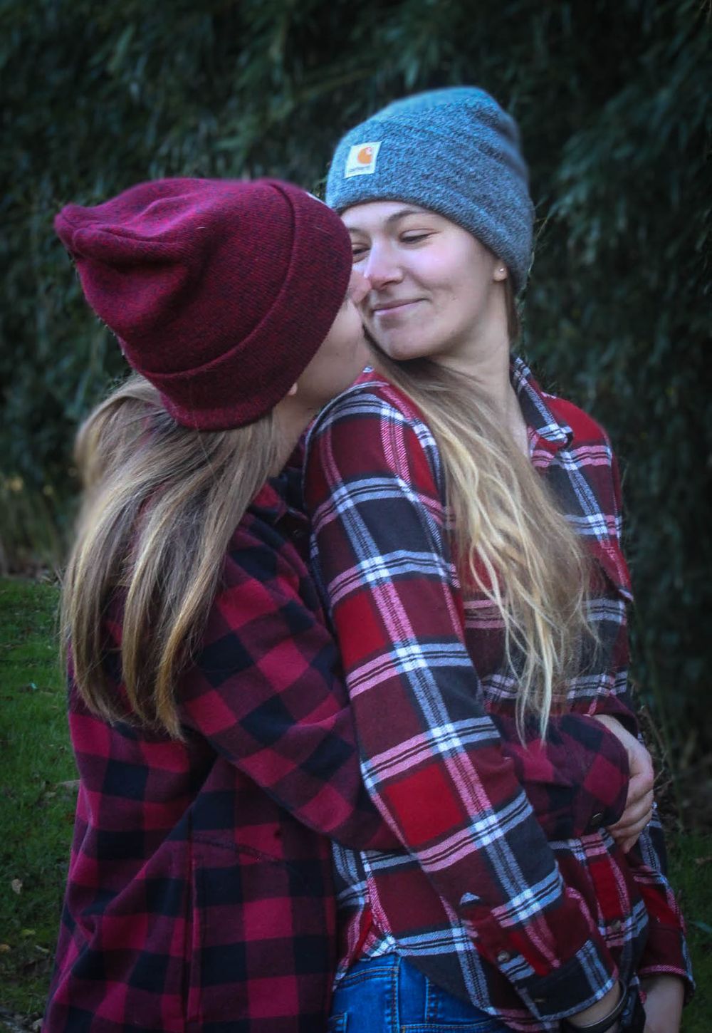 Two people in plaid flannel shirts and winter hats embrace outdoors.