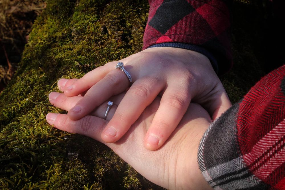 Close-up of hands with engagement ring resting on mossy surface.