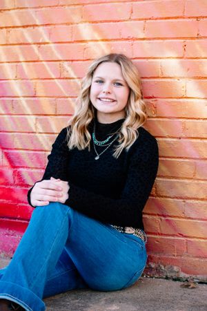 Person in black turtleneck and jeans sitting against orange brick wall with necklace accessories.