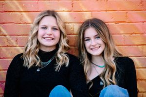 Two friends in matching black sweaters pose together against colorful brick wall background.