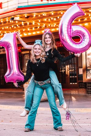 Two friends celebrating with large pink number 16 balloons outside a venue with twinkling lights.