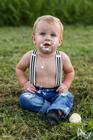 Baby in denim overalls enjoys cake smash photo session on grass.