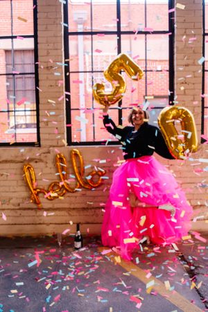 Person in pink tulle skirt celebrates with gold balloons and confetti in industrial space.