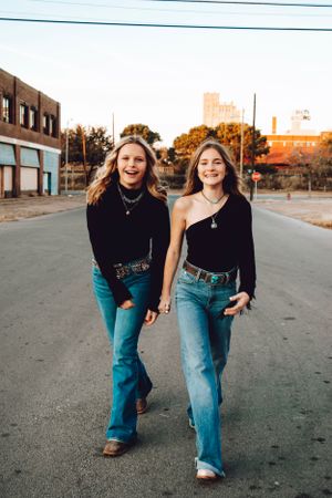 Two friends in black tops and blue jeans walking down urban street at sunset.