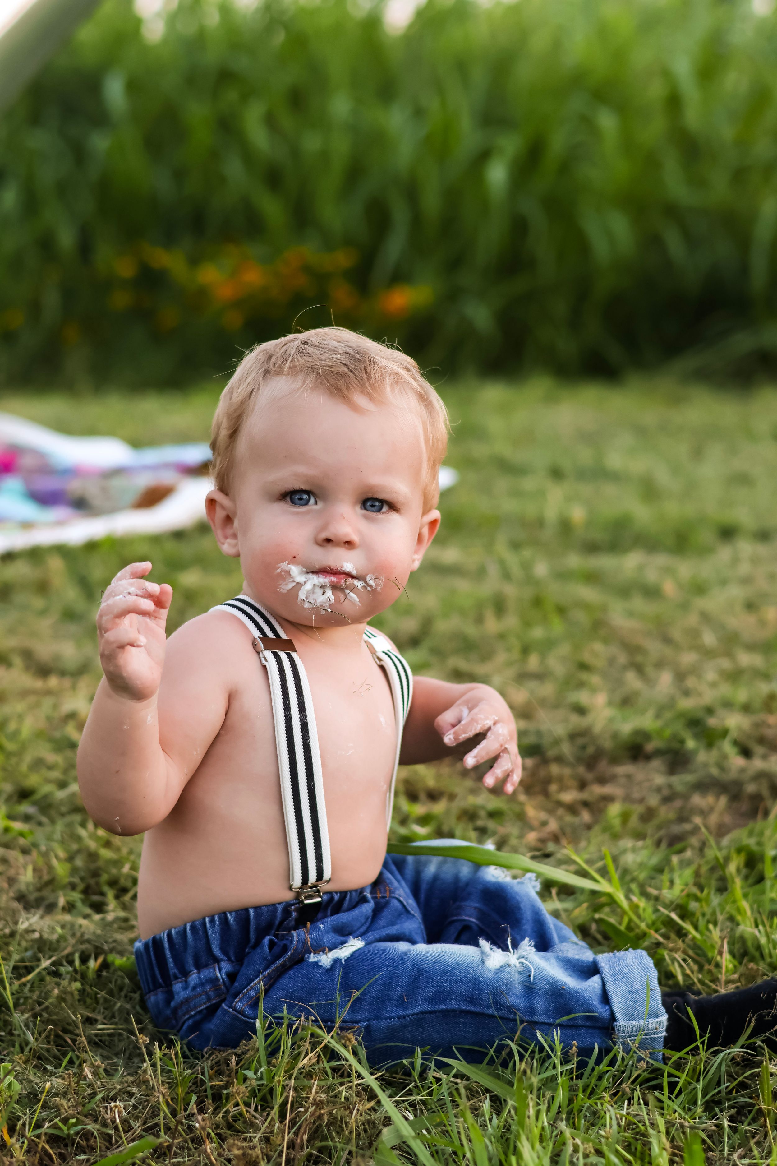 Baby in suspenders plays on grass during outdoor photo session.