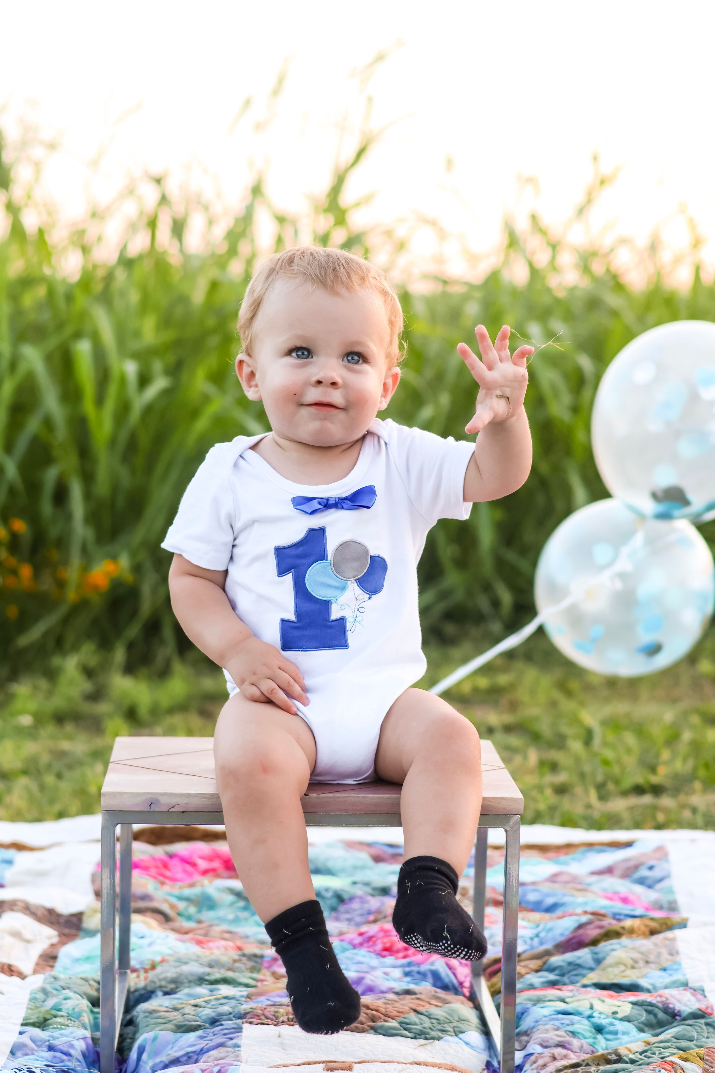 Baby in formal wear with blue bow tie sits on colorful blanket with balloons.