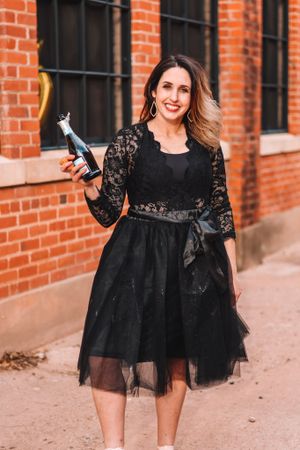 Woman in black lace dress posing with champagne bottle against brick wall.