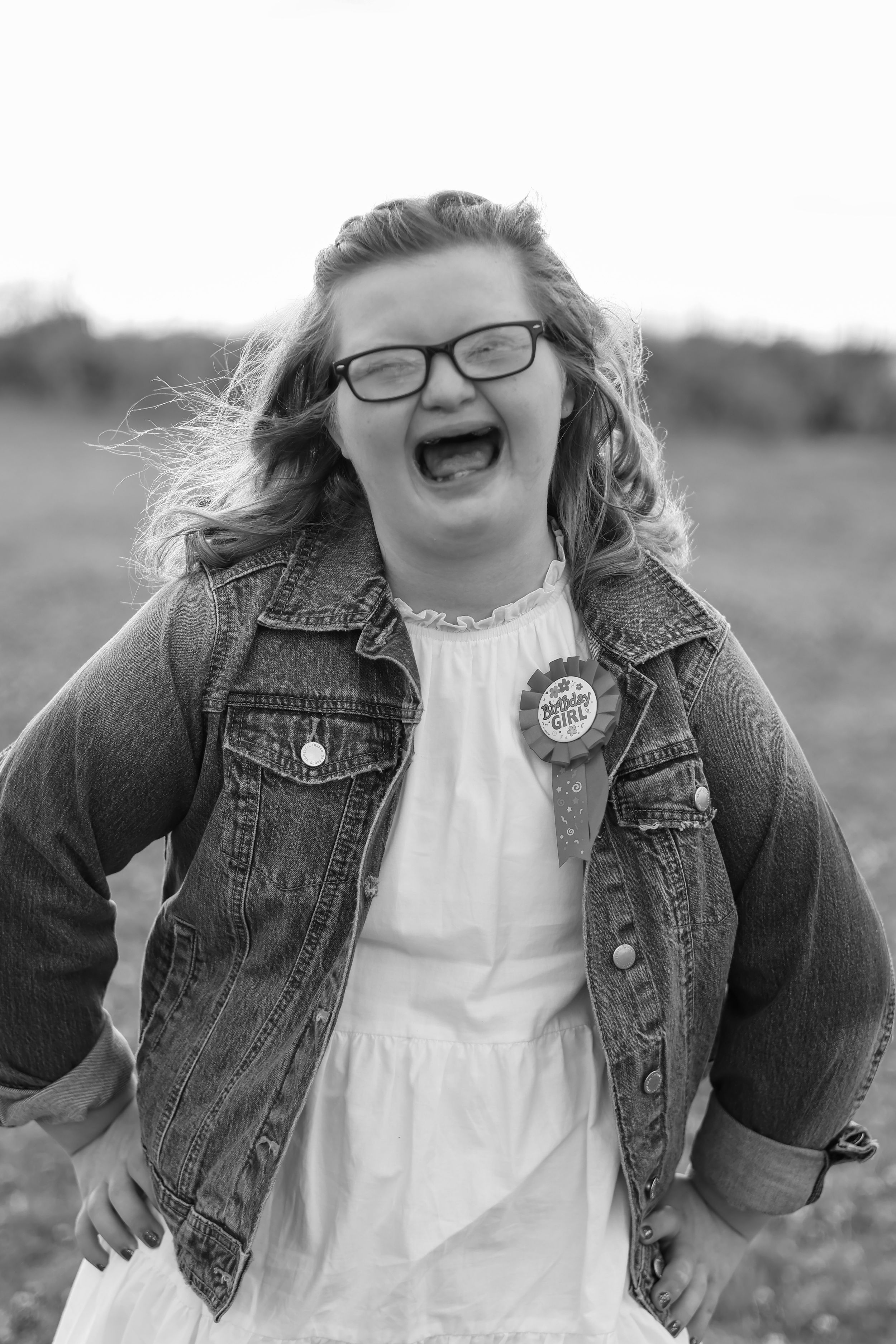 Person wearing denim jacket and white dress posing joyfully outdoors.