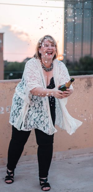 Person in white lace top celebrates with champagne spray at sunset.