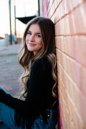 Portrait of a person in black sweater leaning against pink brick wall with long wavy hair.