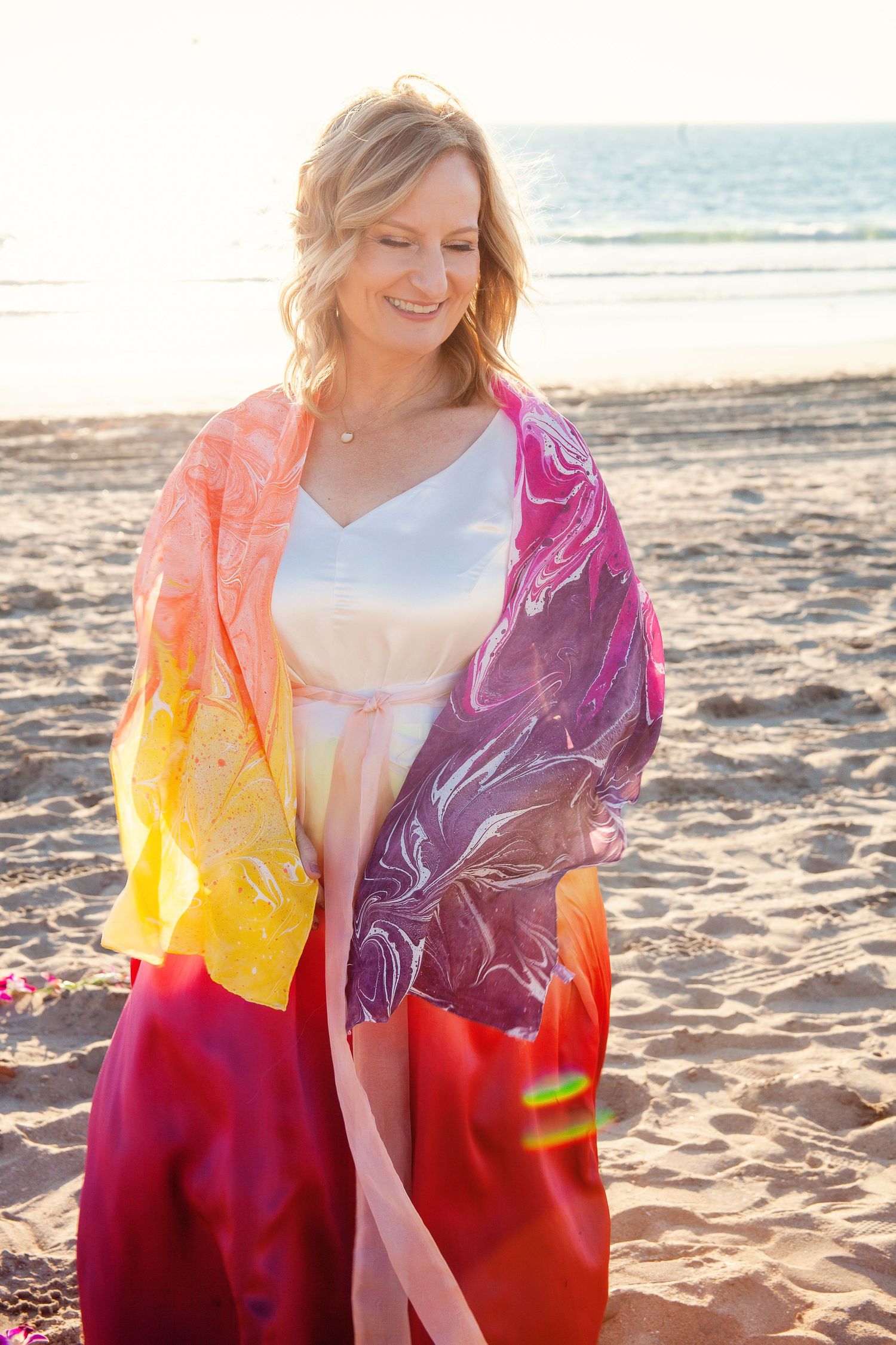 Person on beach wrapped in colorful tie-dye shawl at sunset.
