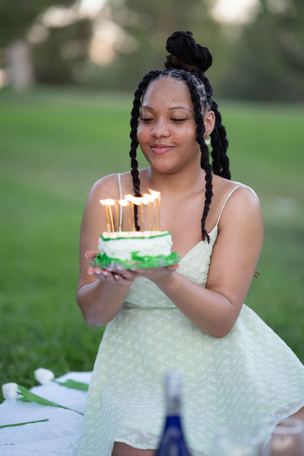 A person in a light green dress holds a small birthday cake with lit candles outdoors.