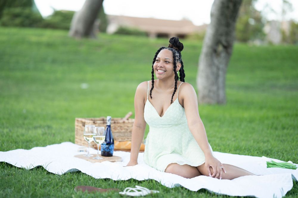 Person relaxing on white blanket during outdoor picnic with drinks and snacks.