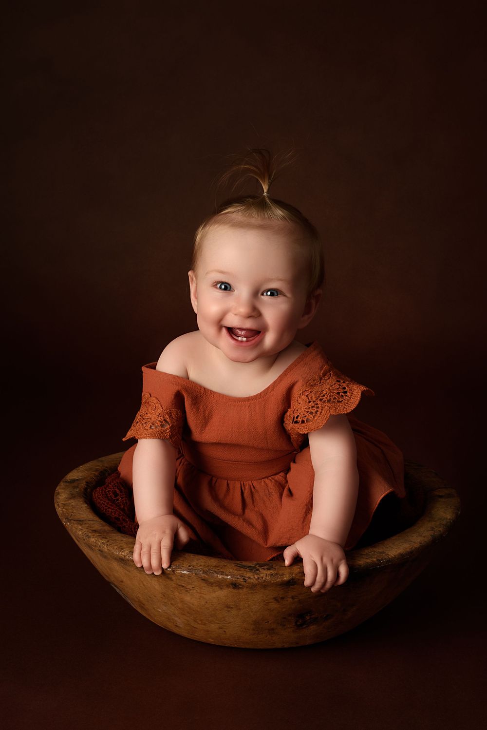 Smiling baby wearing rust-colored dress sitting in wooden bowl against dark background.
