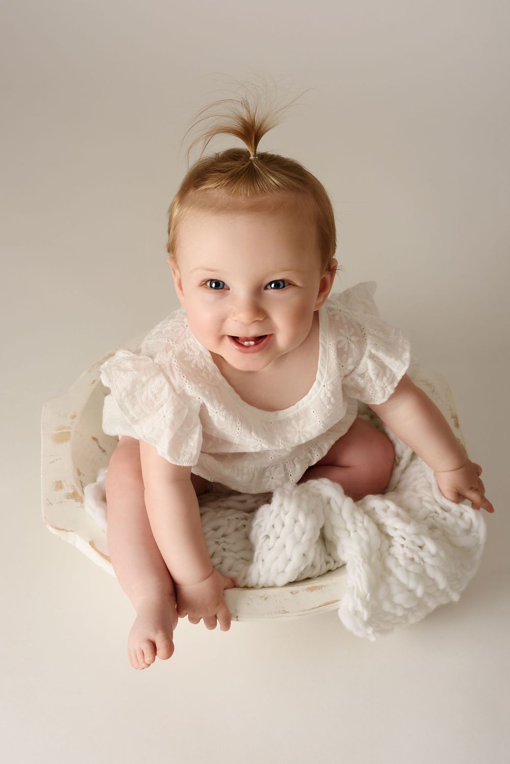 Baby in white lace dress sitting in a white bowl with white, knitted layer with joyful expression.