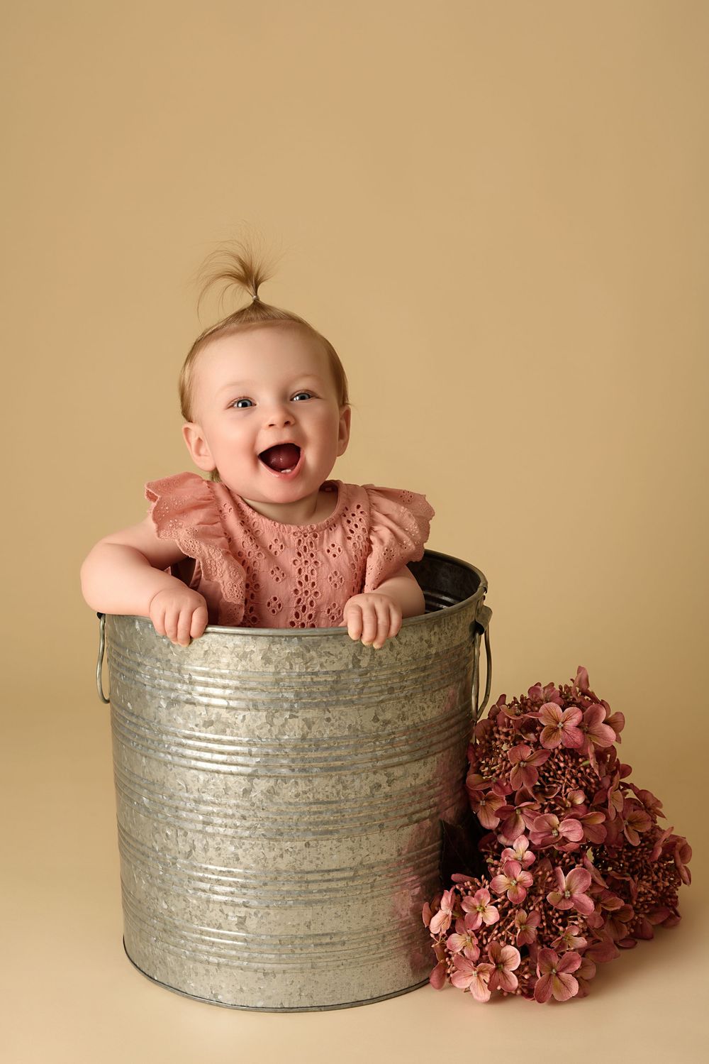 Happy baby in pink dress sitting in metal bucket with flowers.