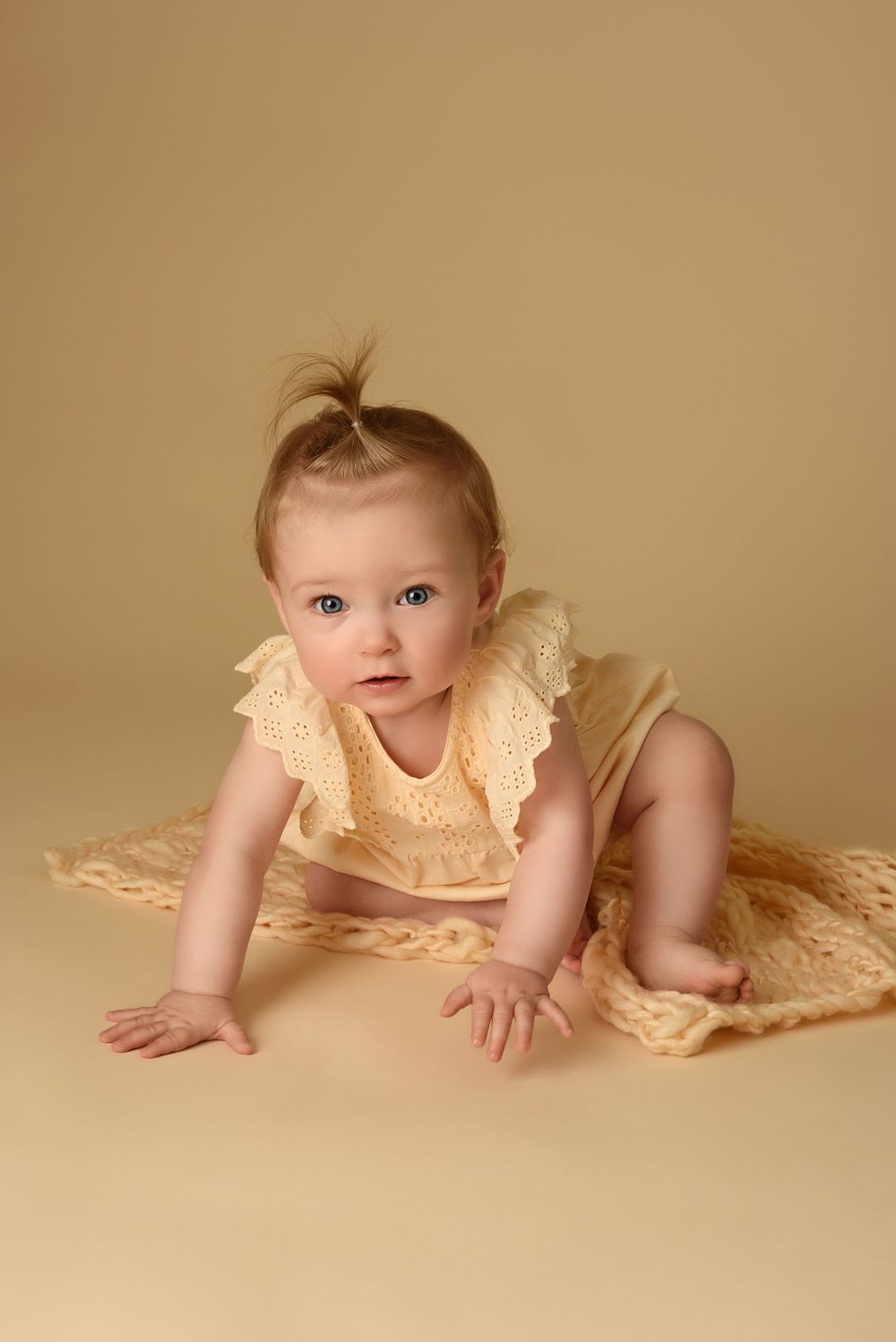 Baby in yellow lace dress crawling on beige background.