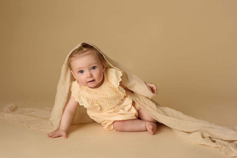 Baby in yellow outfit with fabric draped overhead on neutral background.