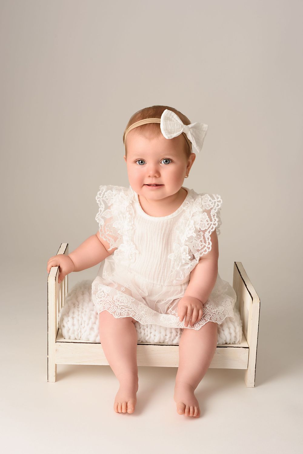 Baby in white lace romper sitting on small white bed.