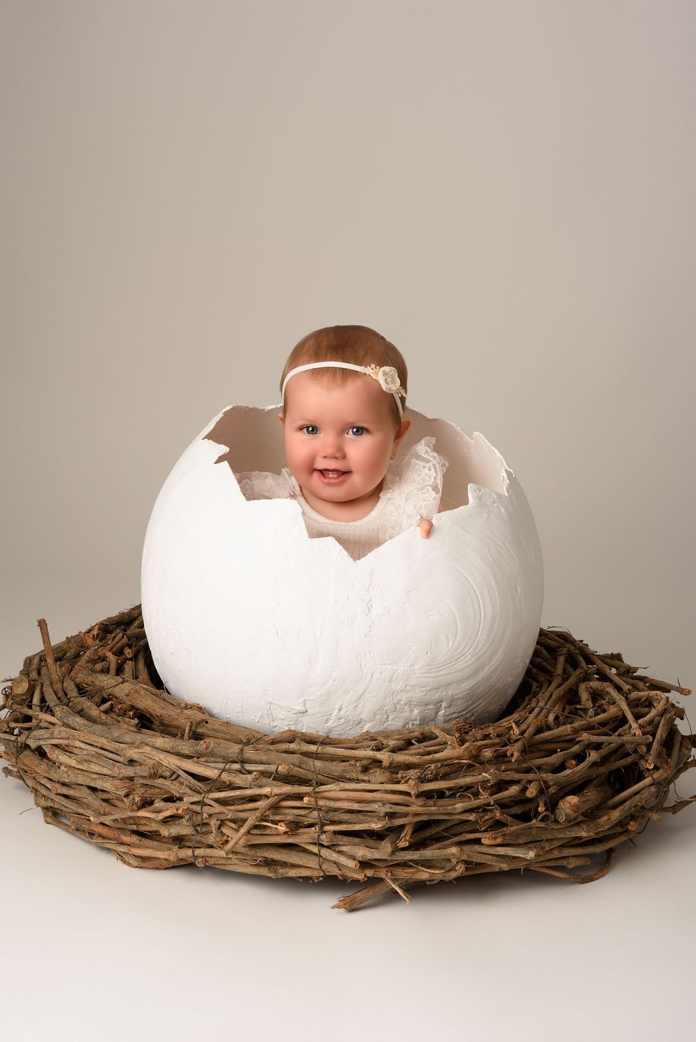 Baby in white dress emerging from large egg prop in nest setting.