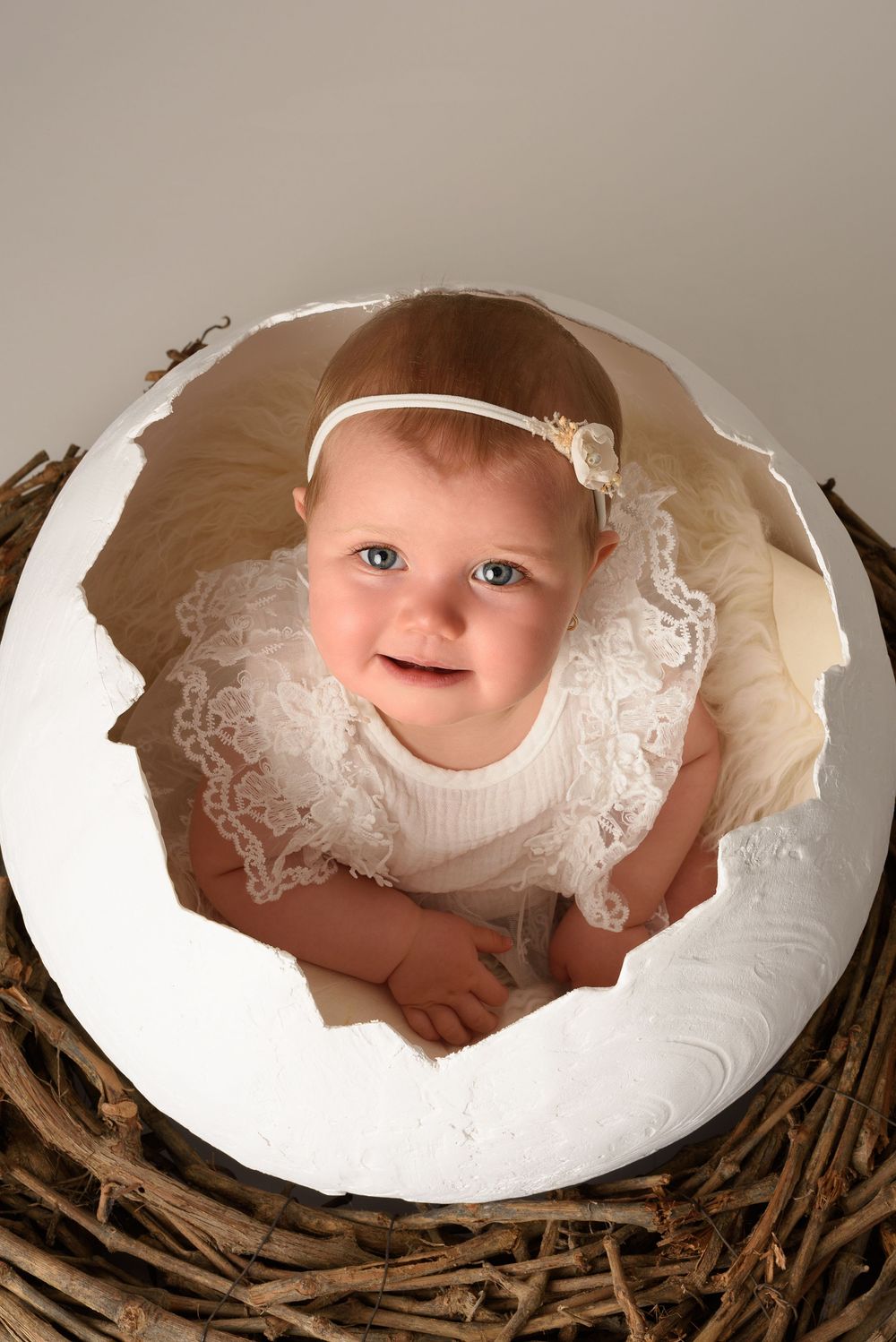 Baby wearing lace dress inside white egg prop nestled in twigs.