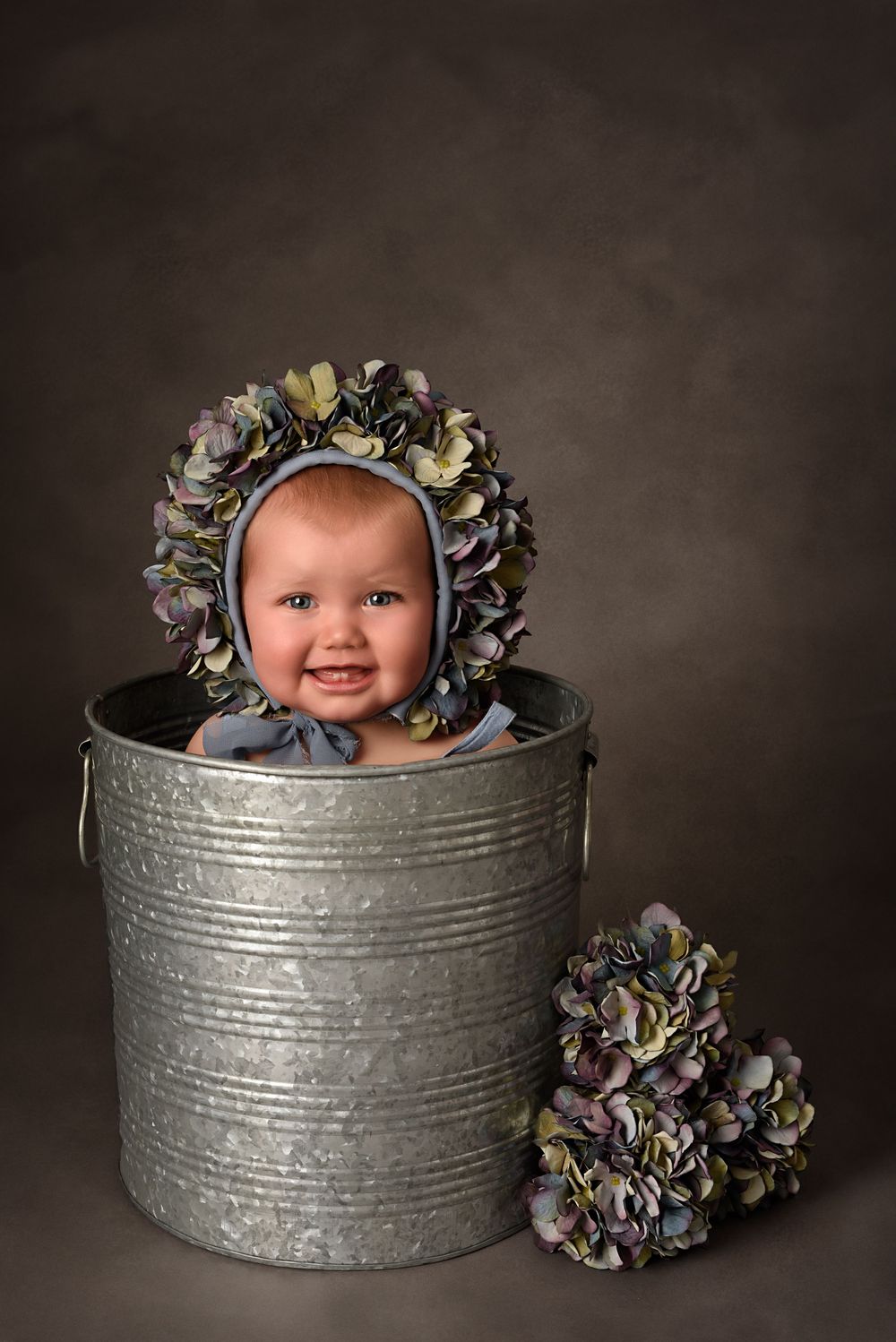 Artistic baby photography setup with flower bonnet vintage metal tub and hydrangea flowers on dark backdrop.