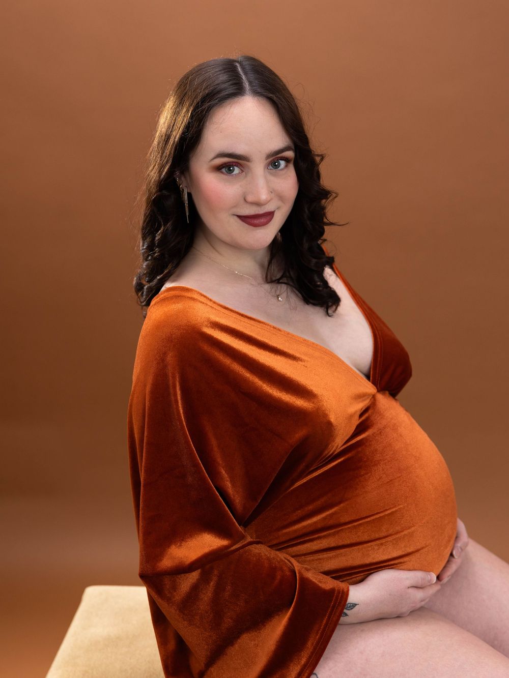 A model in a luxurious burnt orange velvet wrap dress poses elegantly against a brown backdrop.