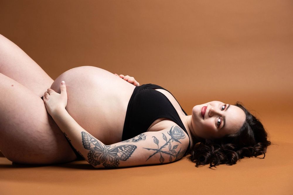 Artistic maternity photo of an expectant mother with tattoos laying on brown backdrop.
