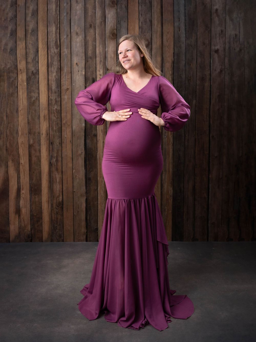 A person in a flowing burgundy maxi dress with long sleeves poses gracefully against a dark wooden wall background.