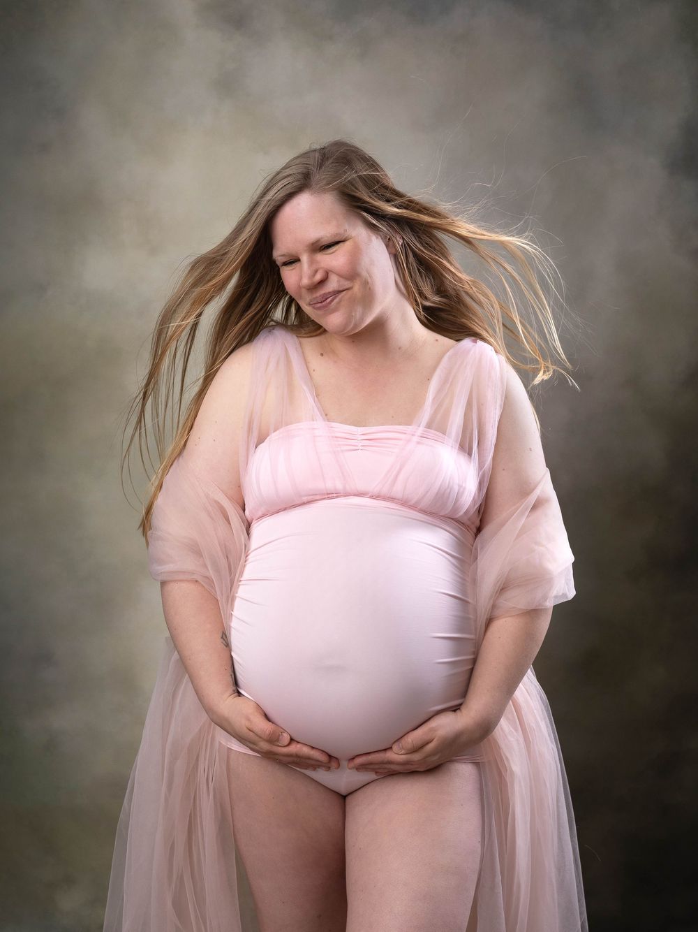 A maternity photoshoot series showing a figure in a pink flowing dress and white leotard against a dark background.