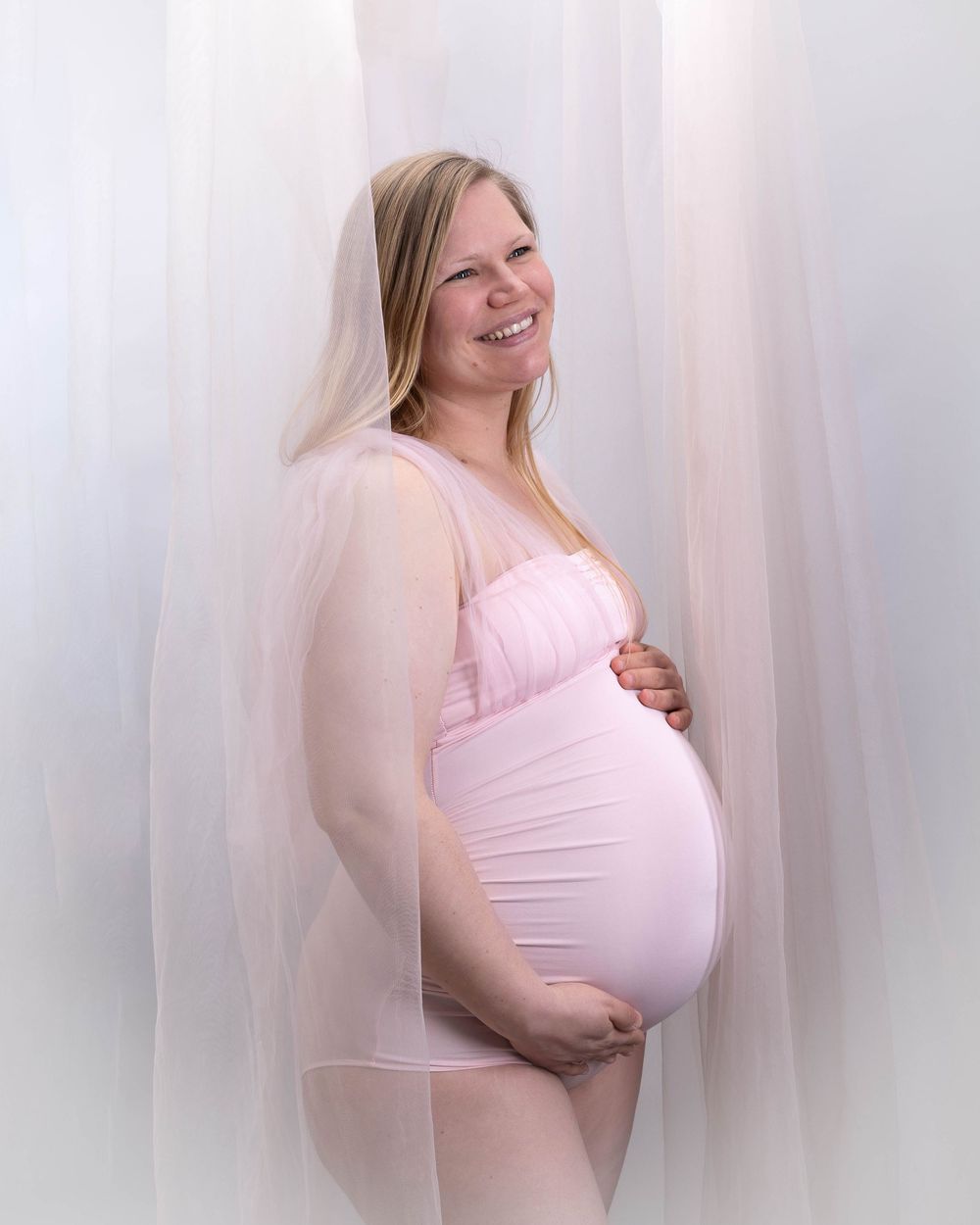 A pregnant woman in a pink dress poses against a white backdrop with soft ethereal lighting.