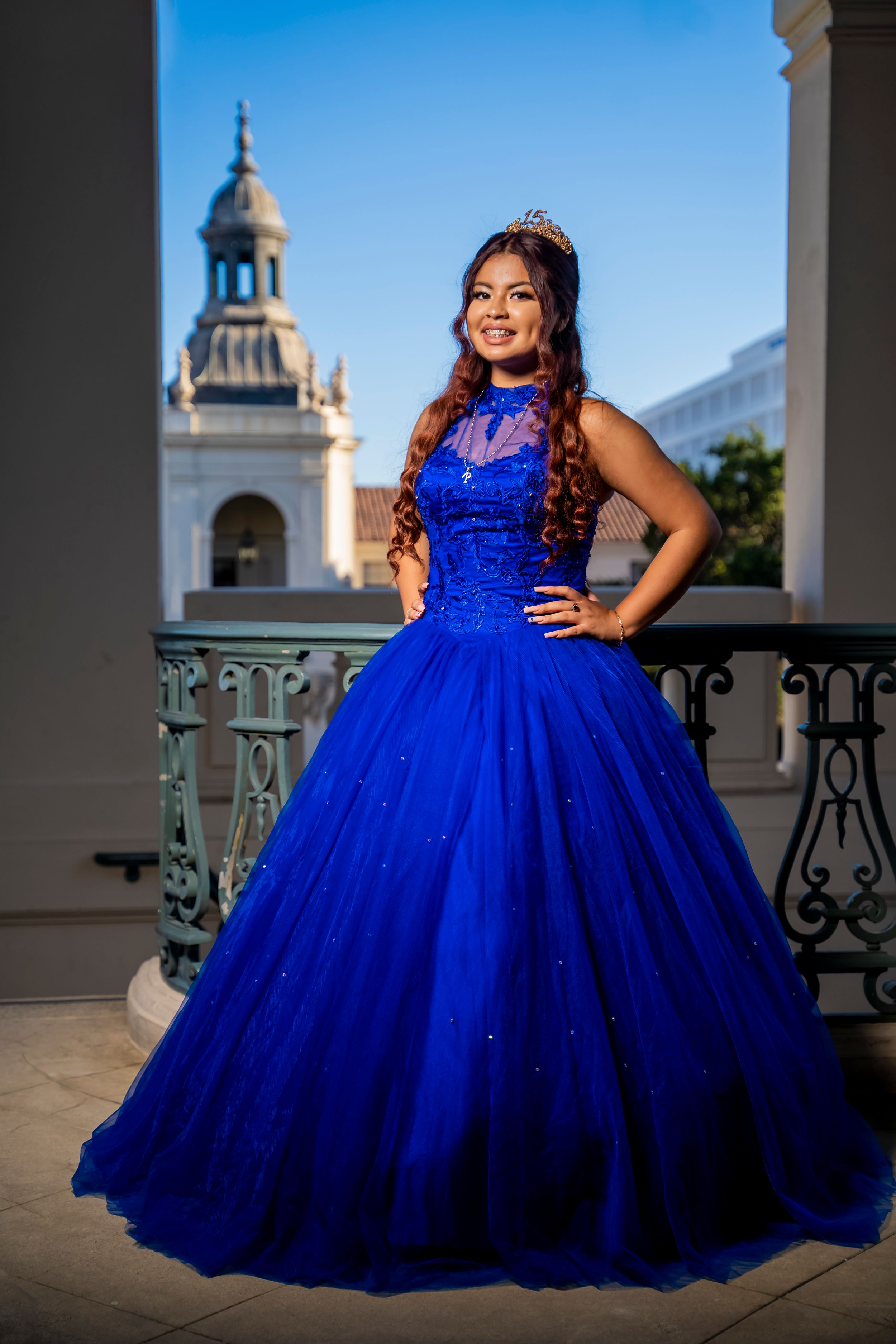 A sparkling royal blue ball gown captured on a balcony with Spanish colonial architecture in the background.
