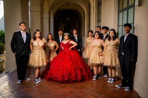 Quinceañera celebration group photo with gold dresses and red gown under arched colonnade.