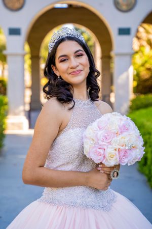 A quinceañera celebration portrait featuring a pink beaded gown and bouquet of light pink roses.