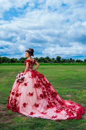 A pink and red floral ball gown with dramatic train photographed in an open field under cloudy skies.