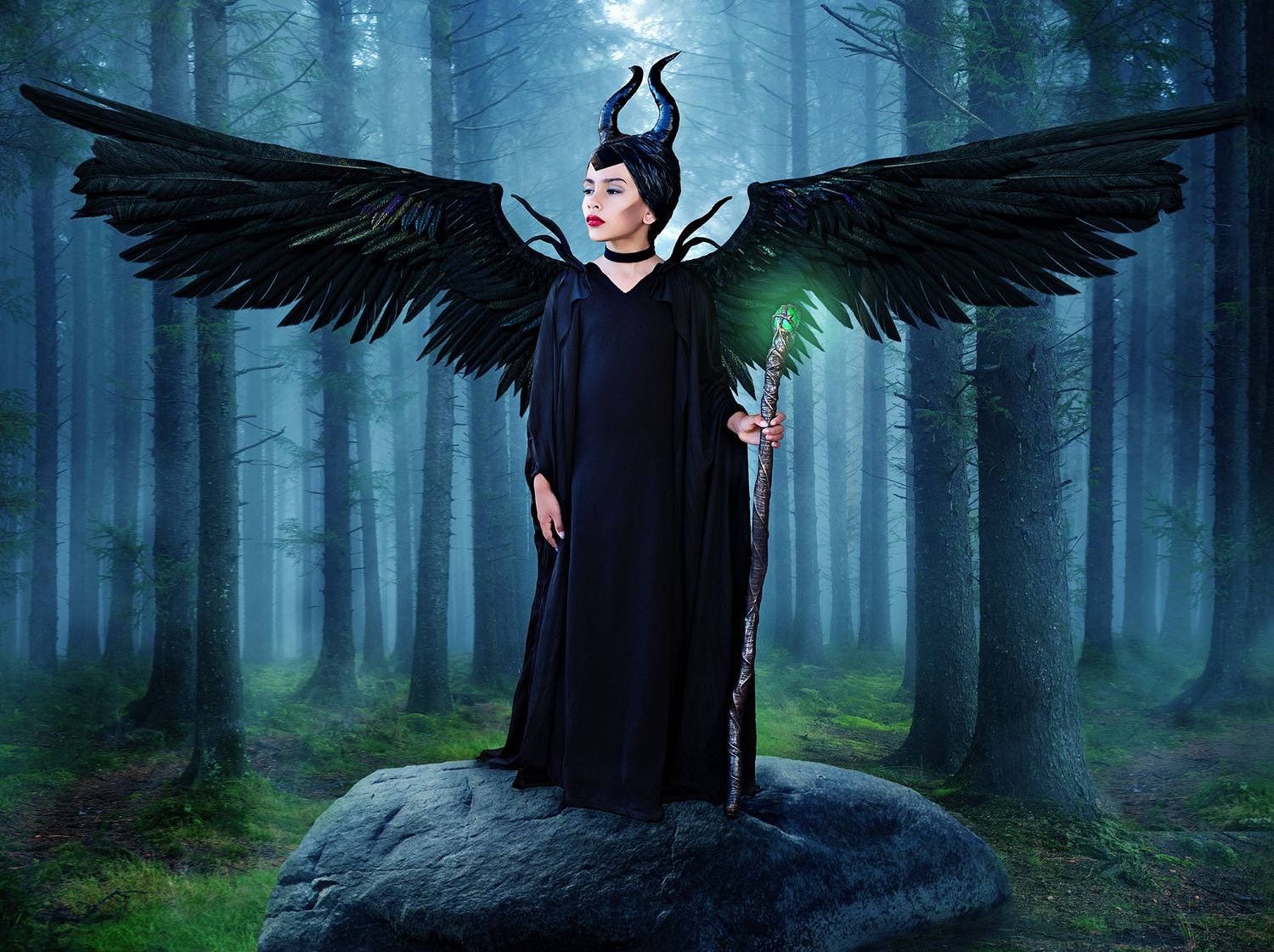 Child dressed as Disney Character Maleficent with wings and glowing staff standing on a boulder in a menacing forest.