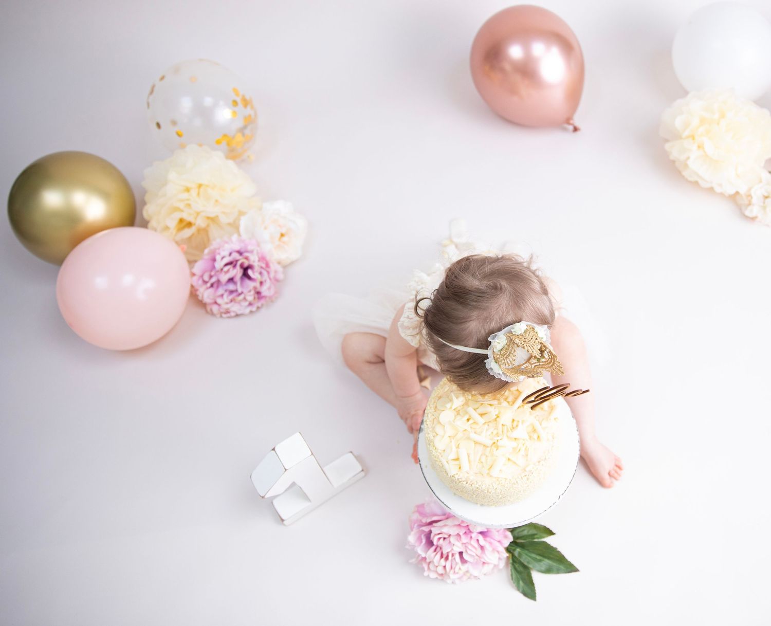 1st birthday cake smash, overhead view of baby eating cake with gold balloons around and pom poms and flowers