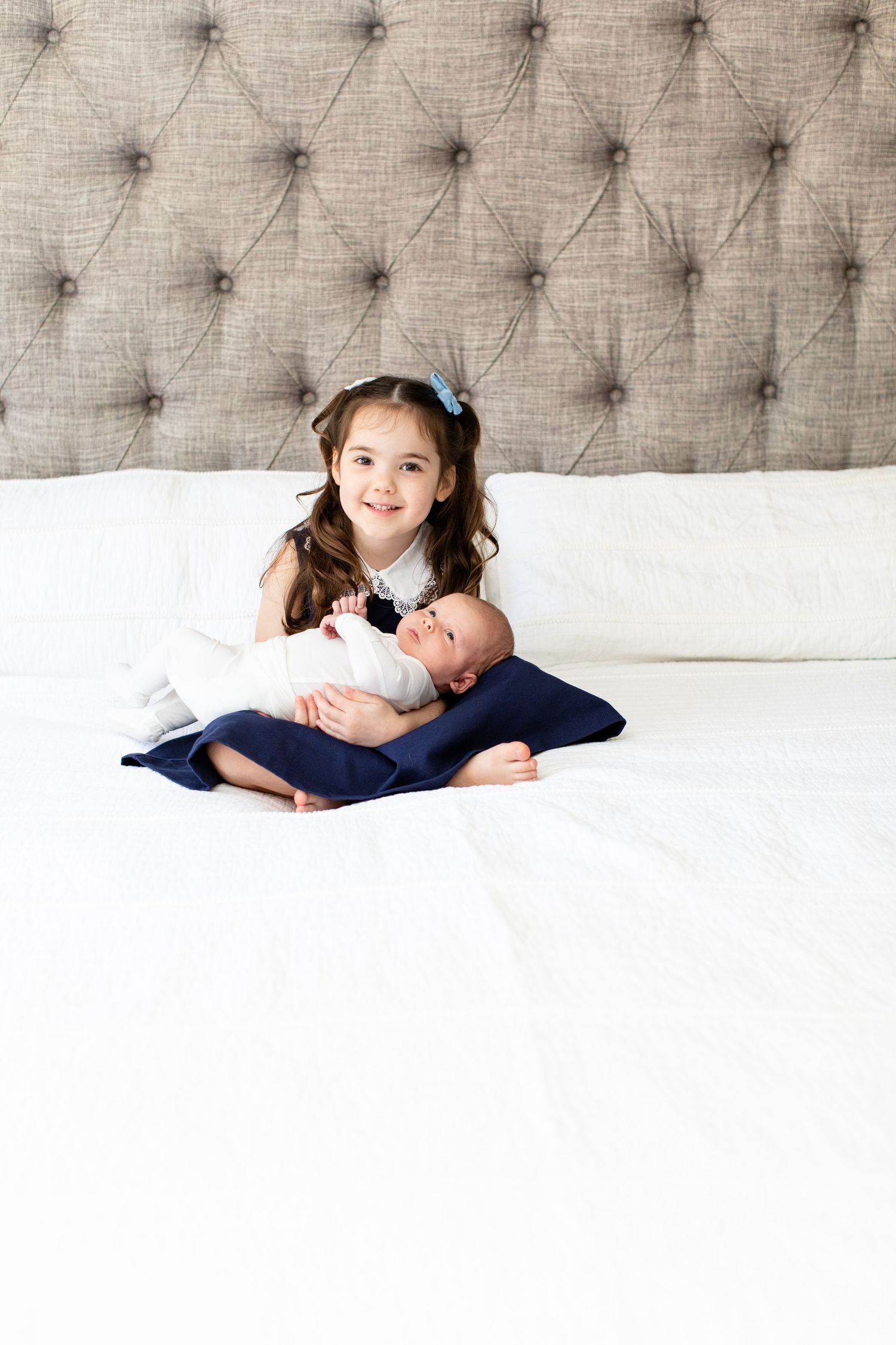 Little girl with hair bows sits with newborn baby on white bedding.