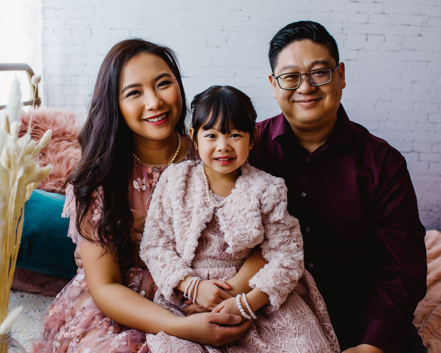 Family of three wearing pink and burgundy outfits sitting together against white brick wall.