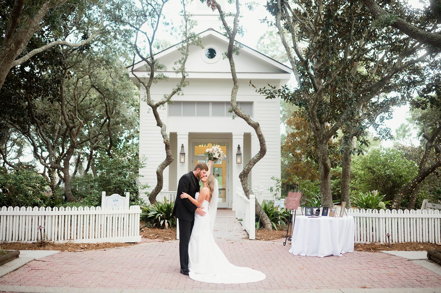 Wedding couple posing in front of white chapel with picket fence and reception table.