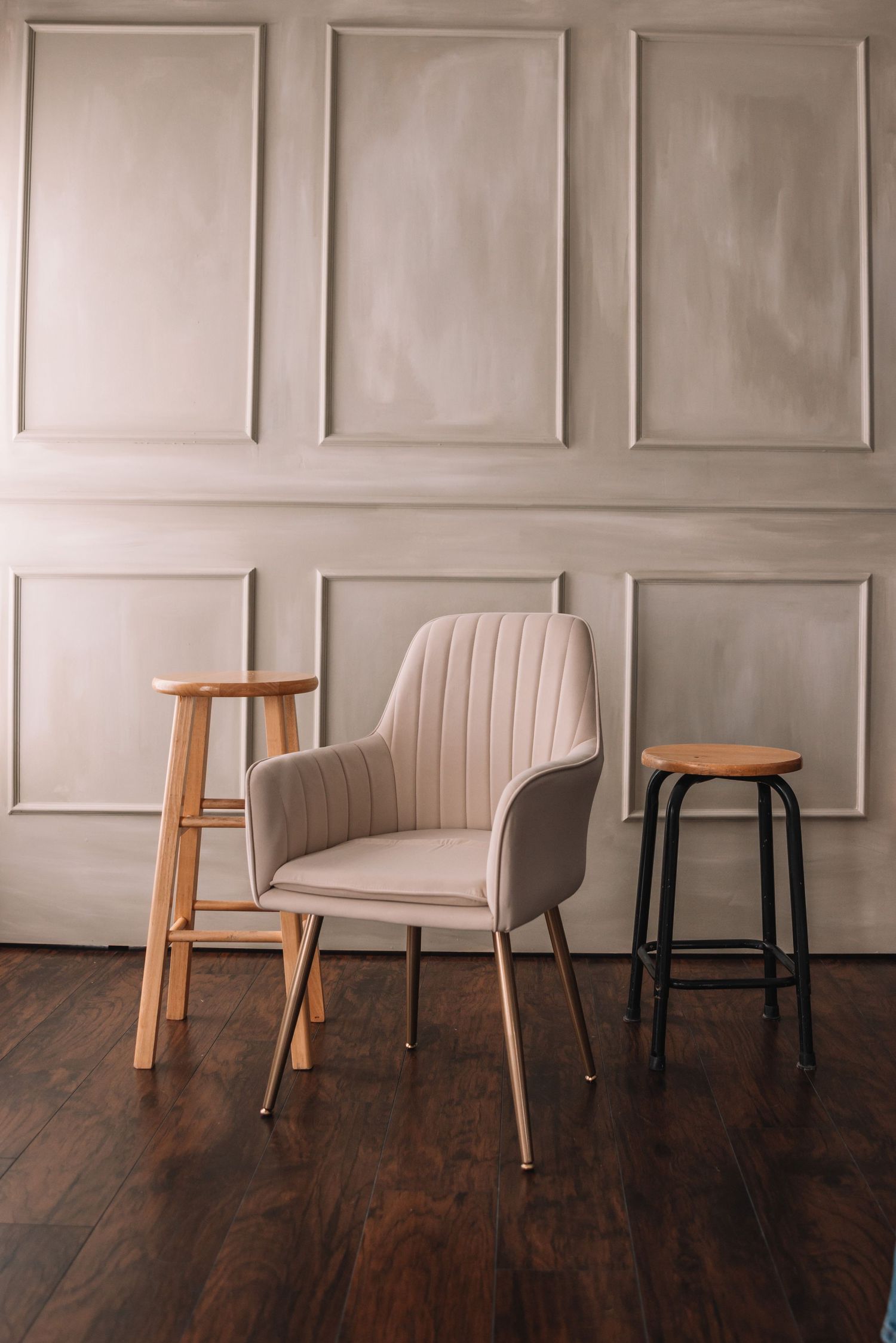 Modern interior setting with beige upholstered chair and wooden stools against paneled wall and hardwood floor.