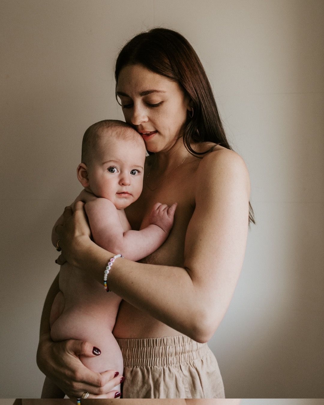 Mother holding chubby baby against neutral wall in warm light.