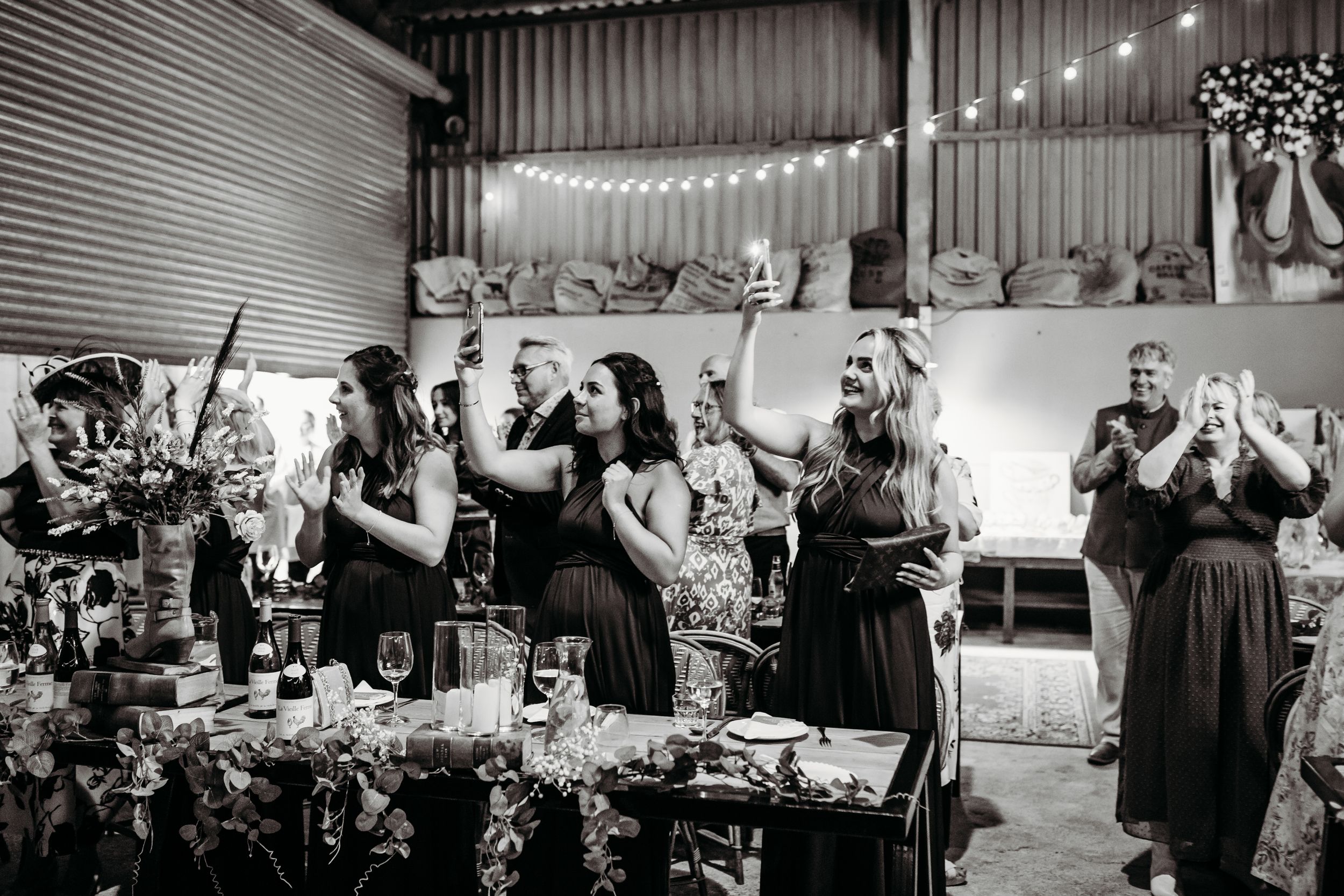 Black and white photo of wedding reception guests celebrating in a rustic venue.