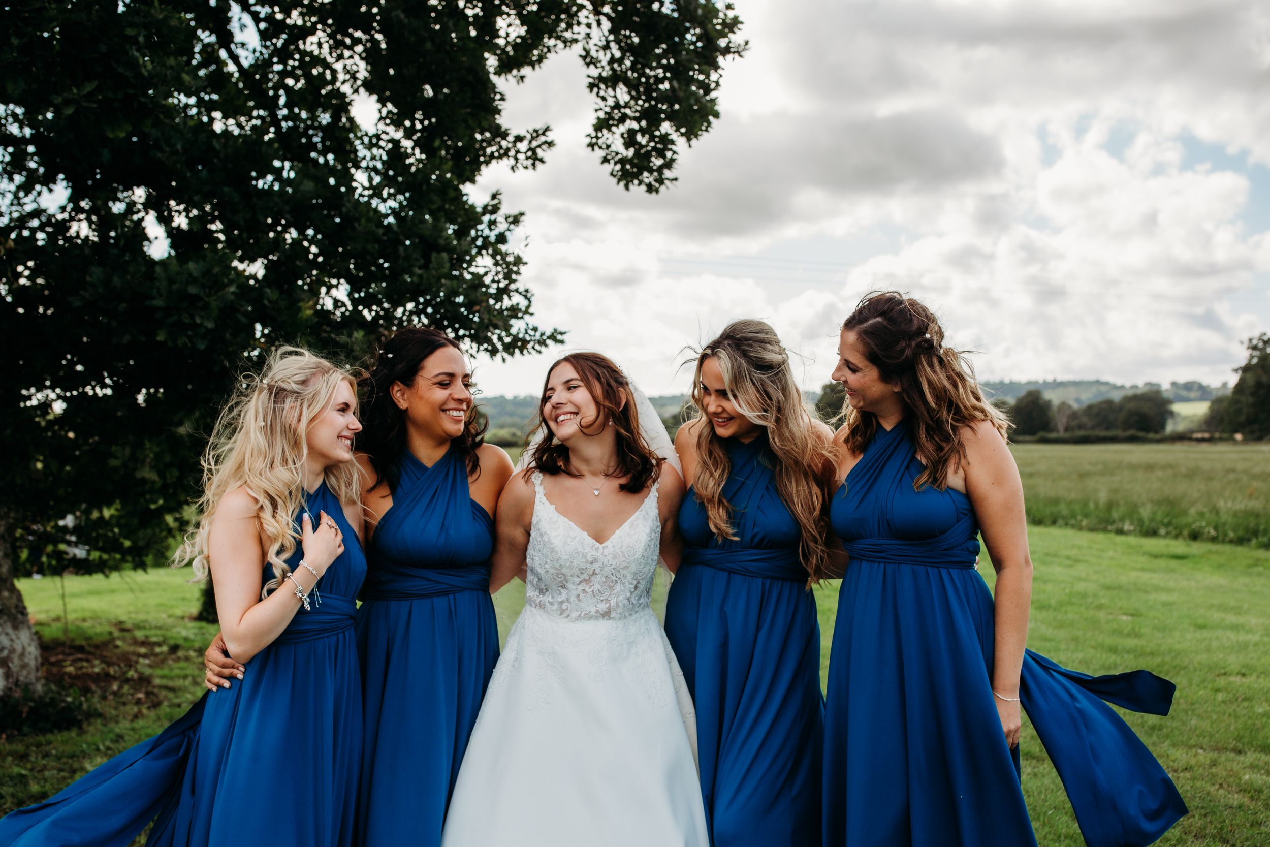 Candid moment of bridesmaids in blue dresses laughing with bride in white gown.